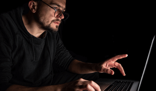 Um homem de óculos trabalha em um laptop no escuro